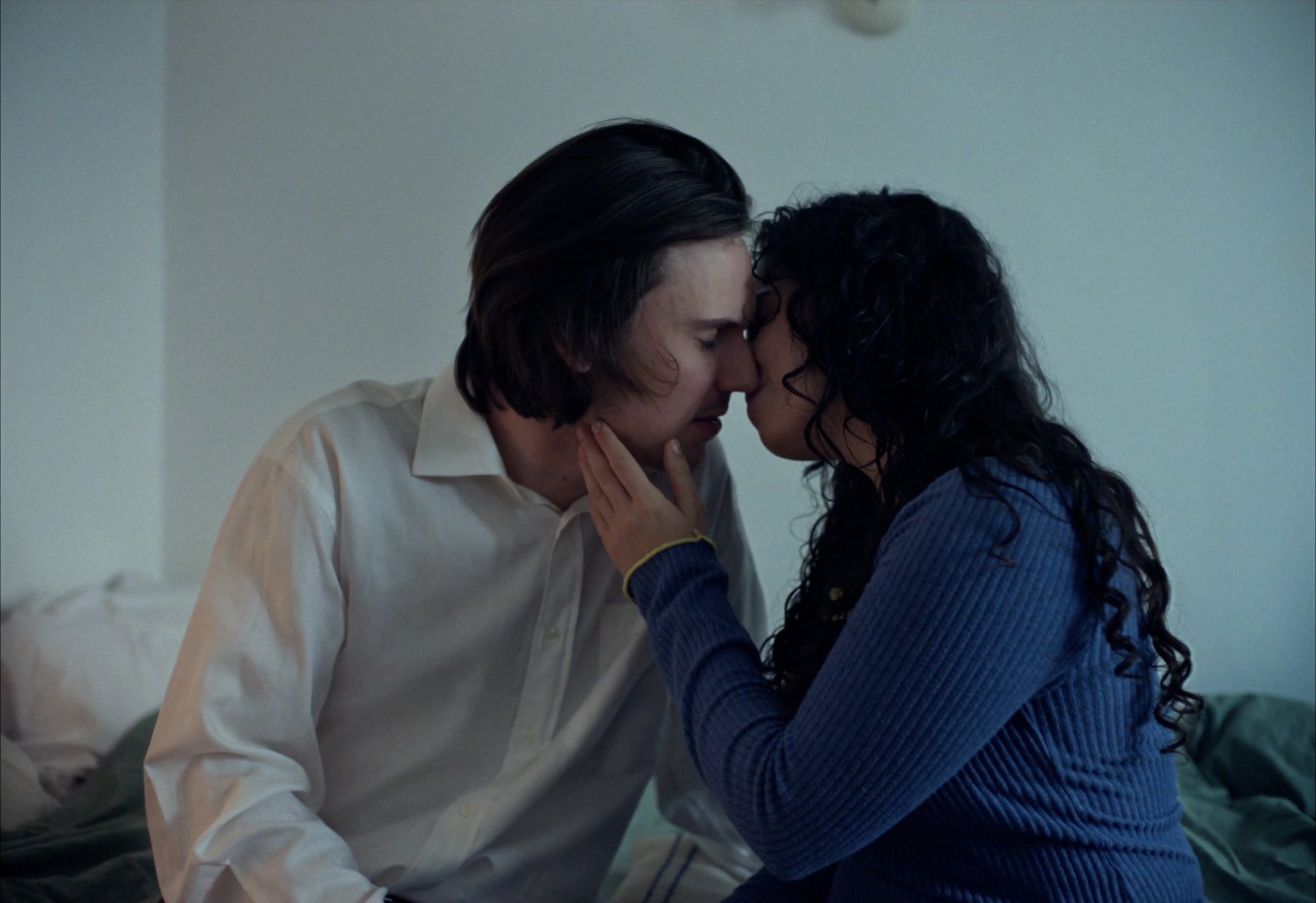 Yasmin and Joseph leaning into each other on a bed, her hand at his jaw.
